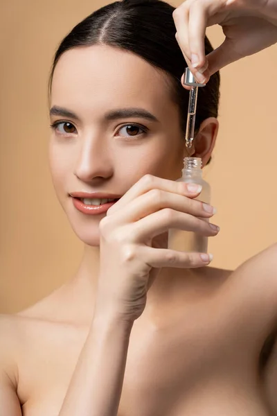 Portrait of confident and smiling asian woman with naked shoulders holding serum isolated on beige — Stock Photo