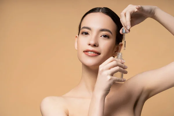 Positive asian woman with naked shoulders posing with cosmetic serum isolated on beige — Stock Photo