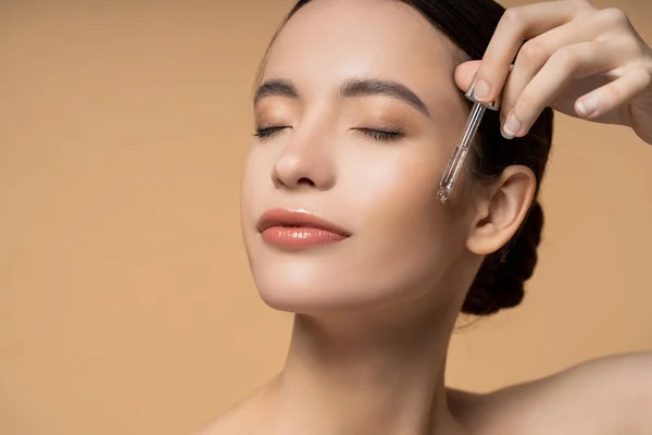 Young asian woman with closed eyes applying cosmetic serum while posing isolated on beige — Stock Photo