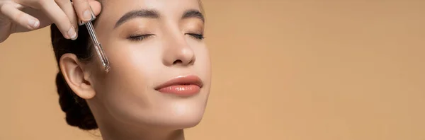 Mujer asiática bastante joven con los ojos cerrados aplicando suero cosmético aislado en beige, pancarta - foto de stock