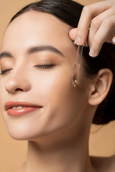 Close up view of cheerful young asian model applying cosmetic serum isolated on beige — Stock Photo