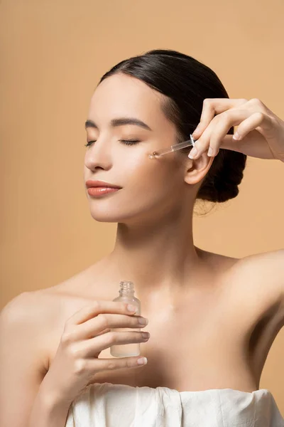 Jolie femme asiatique avec les yeux fermés en haut appliquant sérum cosmétique tout en posant isolé sur beige — Photo de stock