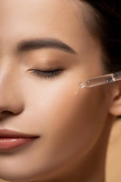Ausgeschnittene Ansicht einer jungen asiatischen Frau, die Kosmetikserum auf die Wange aufträgt, während sie isoliert auf beige steht — Stockfoto