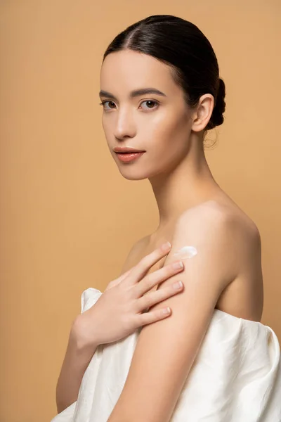 Young brunette asian woman applying body cream on shoulder while standing isolated on beige — Stock Photo