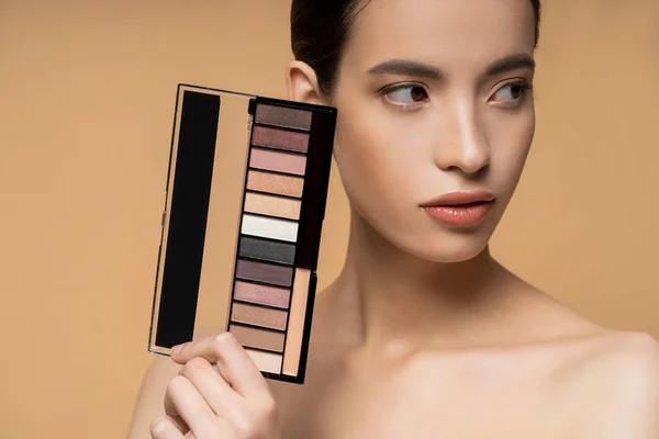Asian woman with naked shoulders holding eyeshadow palette and looking away isolated on beige — Stock Photo