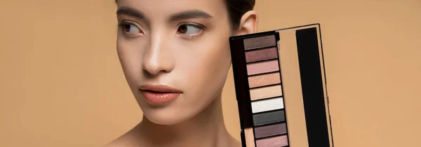 Pretty young asian woman with natural makeup holding eyeshadow palette isolated on beige, banner — Stock Photo