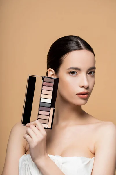 Pretty young asian woman in top with naked shoulders holding eyeshadow palette isolated on beige — Stock Photo