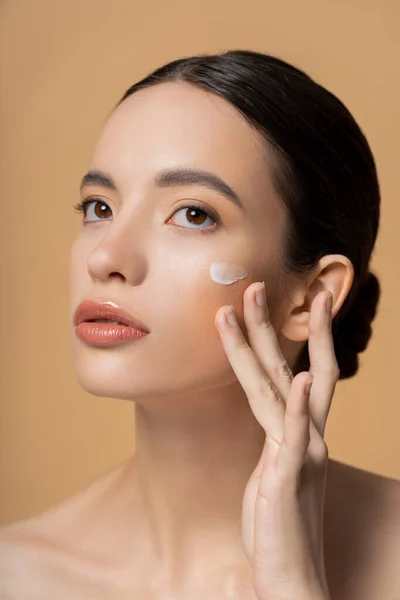 Young asian woman with naked shoulders applying face cream isolated on beige — Stock Photo