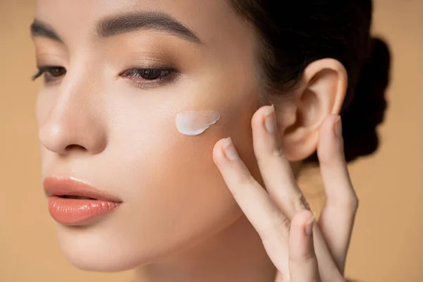 Nahaufnahme einer jungen asiatischen Frau, die Gesichtscreme auf die Wange aufträgt, isoliert auf beige — Stockfoto