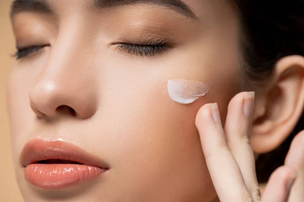 Vista da vicino della giovane donna asiatica con pelle perfetta applicare crema viso sulla guancia isolata sul beige — Foto stock