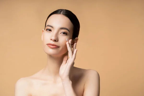 Young asian woman looking at camera while applying face cream and standing isolated on beige — Stock Photo