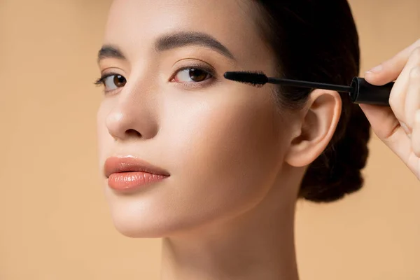 Close up view of young asian woman applying mascara and looking at camera isolated on beige — Stock Photo