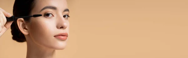 Schöne asiatische Frau, die Wimperntusche aufträgt und isoliert auf beige, Banner in die Kamera schaut — Stockfoto