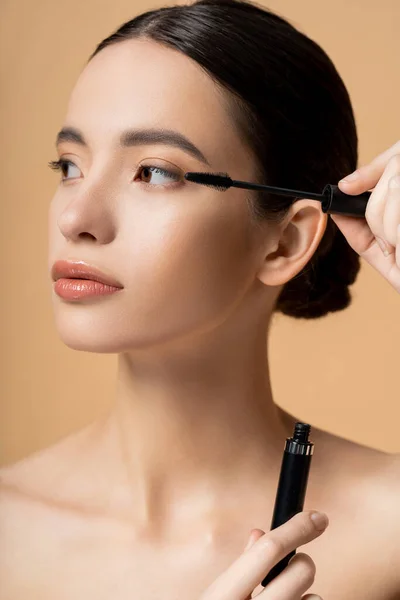Young asian woman with natural makeup holding mascara and applicator isolated on beige — Stock Photo