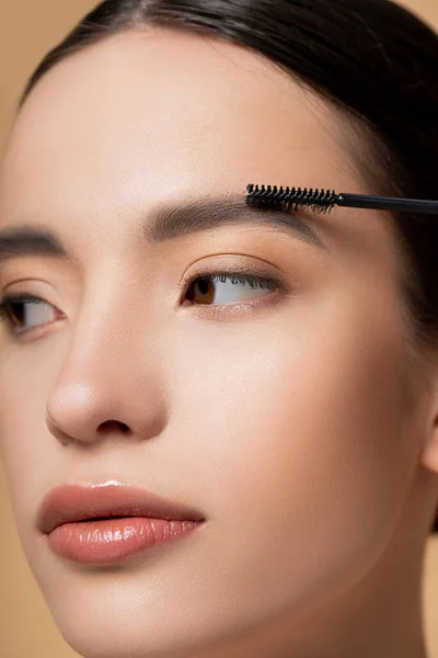 Close up view of young asian woman with natural makeup applying eyebrow gel isolated on beige — Stock Photo