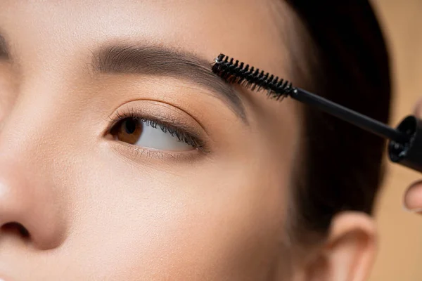 Close up view of young asian woman applying eyebrow gel while doing makeup isolated on beige — Stock Photo