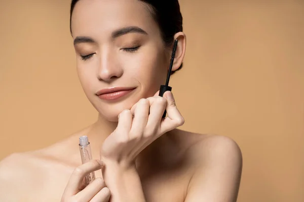 Young asian woman with naked shoulders and closed eyes holding mascara isolated on beige — Stock Photo