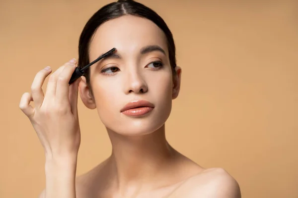 Young brunette asian woman with naked shoulders applying eyebrow gel while posing isolated on beige — Stock Photo