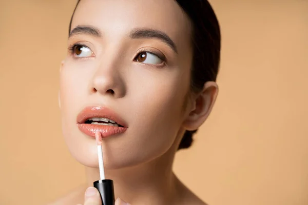 Retrato de joven modelo asiático aplicando brillo de labios y mirando hacia otro lado aislado en beige - foto de stock