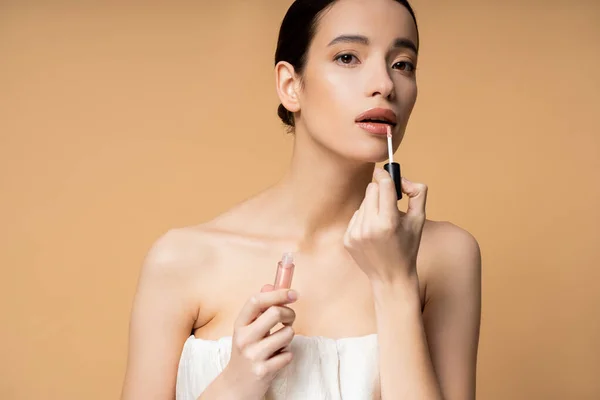 Pretty young asian woman in top looking at camera while applying lip gloss isolated on beige — Stock Photo