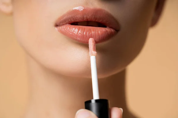 Close up view of young woman applying lip gloss while doing makeup isolated on beige — Stock Photo