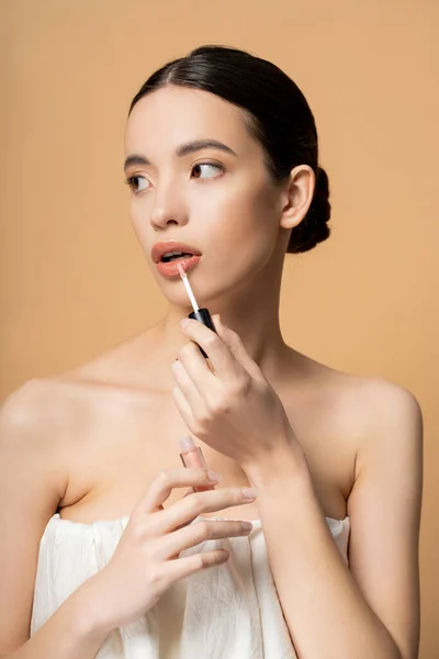 Pretty asian model with naked shoulders in top applying lip gloss and looking away isolated on beige — Stock Photo