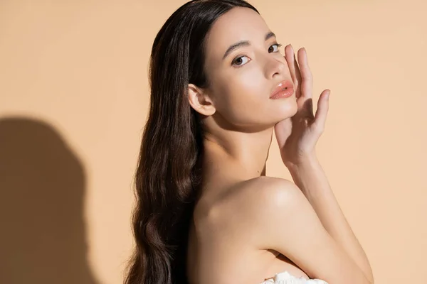 Hübsche asiatische Frau mit langen Haaren und nackter Schulter, die die Wange berührt und auf beigem Hintergrund steht — Stockfoto