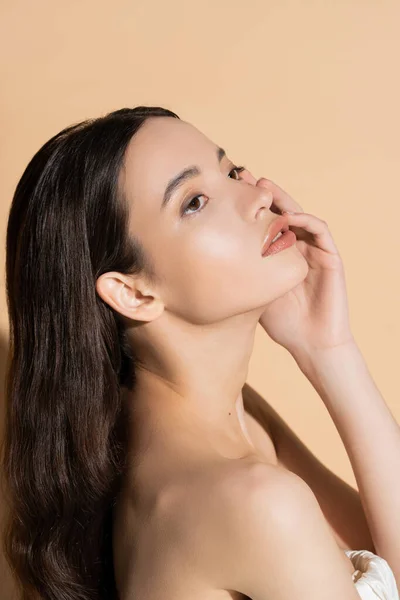 Mulher asiática bonita com maquiagem natural tocando bochecha enquanto está de pé no fundo bege — Fotografia de Stock