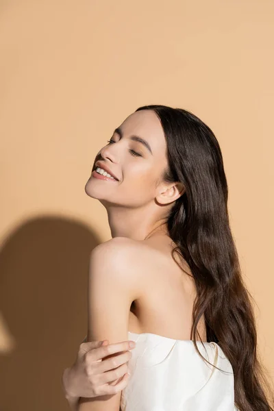 Smiling and long haired asian woman with naked shoulder standing on beige background with shadow — Stock Photo