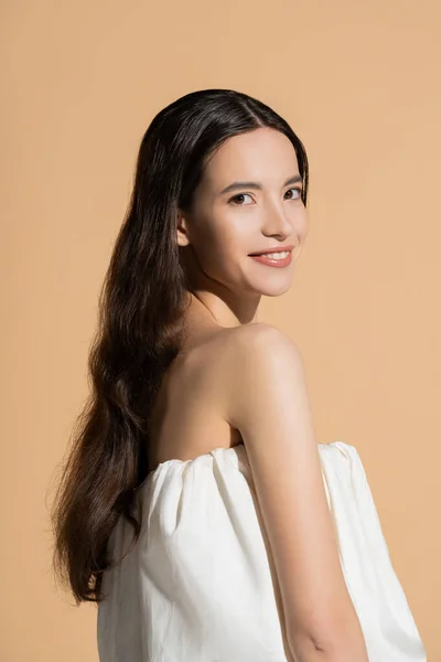 Веселая молодая азиатская модель с длинными волосами, смотрящая в камеру, стоя изолированно на бежевом — стоковое фото
