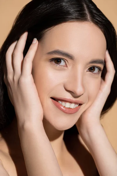 Retrato de modelo asiático positivo con maquillaje natural tocando el cabello aislado en beige - foto de stock