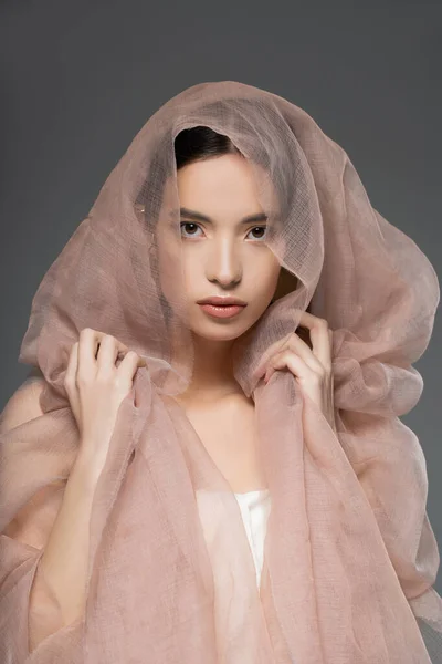 Joven mujer asiática con maquillaje natural tocando tela beige y mirando a la cámara aislada en gris - foto de stock