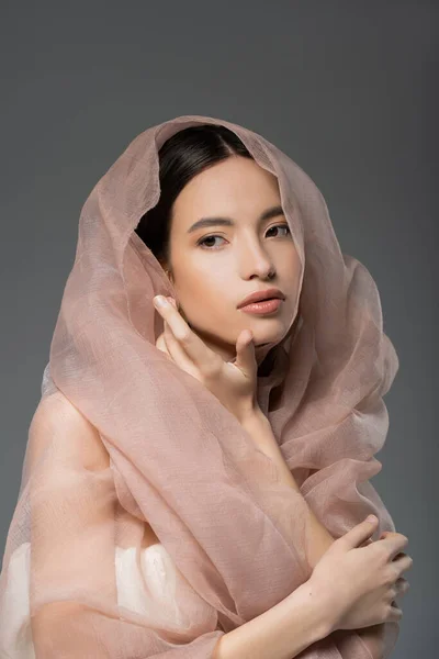 Portrait of beautiful young asian woman posing with beige cloth while standing isolated on grey — Stock Photo
