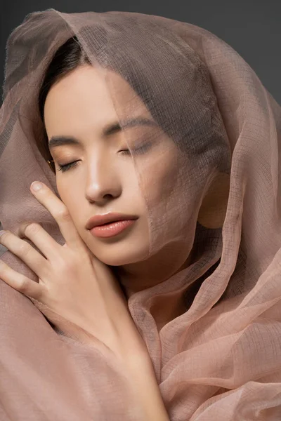 Retrato de hermosa mujer asiática joven con maquillaje natural posando con tela beige aislada en gris - foto de stock