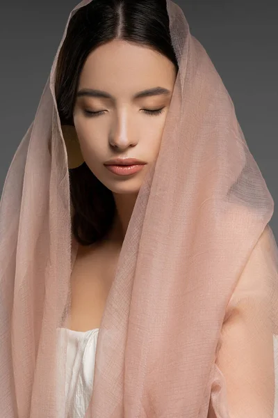 Досить молода азіатська жінка з закритими очима позує в бежевій тканині і стоїть ізольовано на сірому — стокове фото