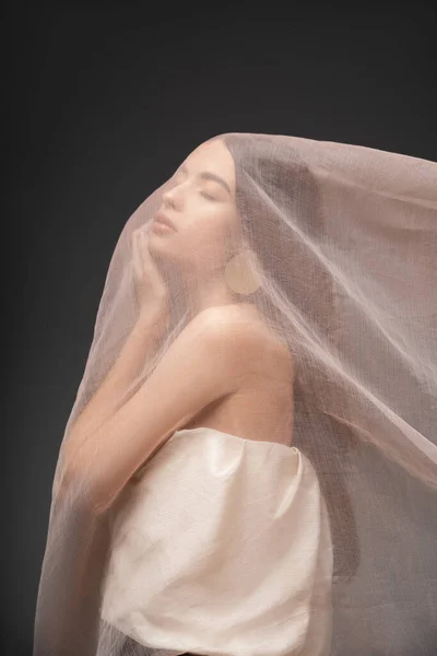 Молода азіатська жінка зверху зворушлива щока, позує під бежевою тканиною ізольовано на сірому — стокове фото