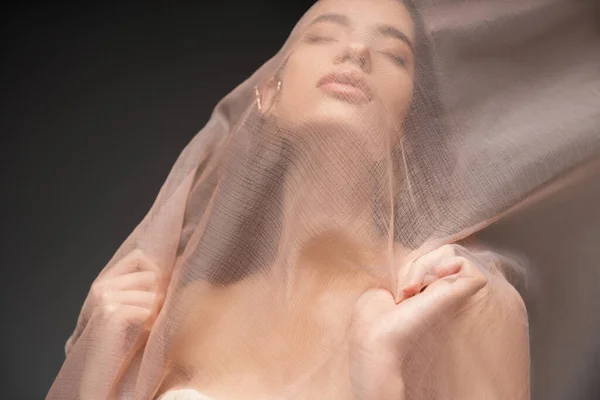 Retrato de modelo asiático con los ojos cerrados posando bajo tela beige mientras está de pie aislado en negro - foto de stock