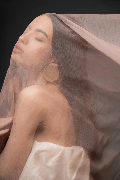 Pretty asian model in top and earring posing under beige cloth while standing isolated on black — Stock Photo