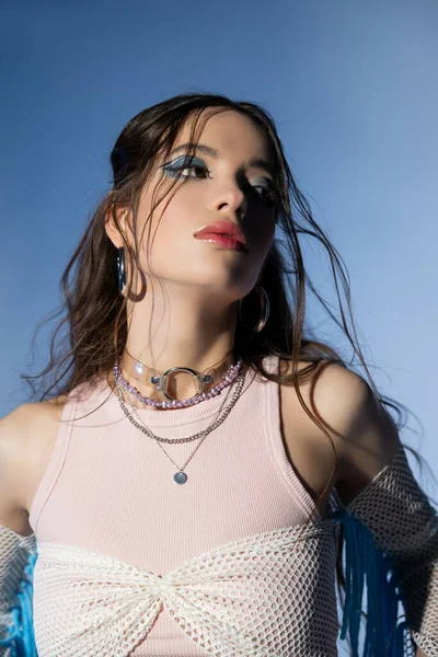 Retrato de modelo asiático com maquiagem ousada e acessórios olhando para longe e posando em fundo azul — Fotografia de Stock