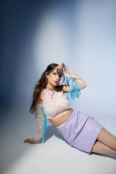 Jeune femme asiatique à la mode en mesh top et jupe en regardant la caméra sur fond bleu — Photo de stock