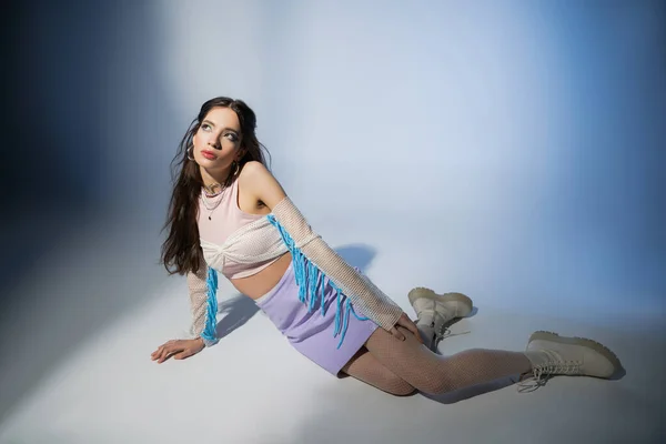 Trendige brünette asiatische Frau in Netztop und Rock schaut weg, während sie auf blauem Hintergrund sitzt — Stockfoto