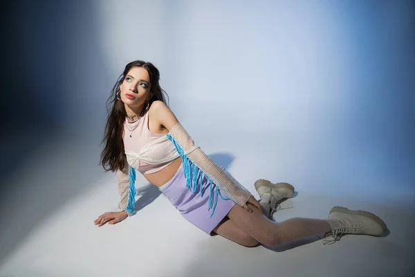 Morena de moda mujer asiática en la parte superior de malla y falda mirando hacia otro lado mientras está sentado sobre fondo azul - foto de stock