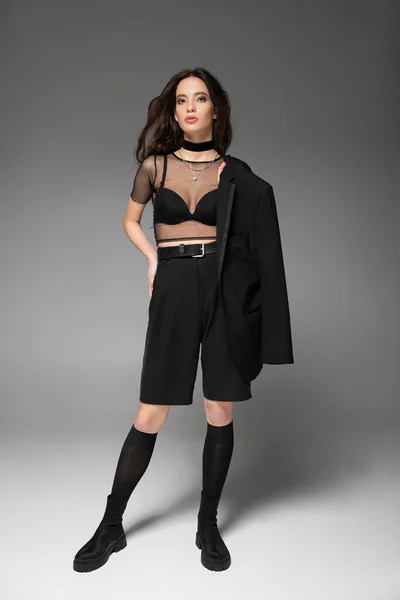 Pleine longueur de femme asiatique à la mode en haut transparent tenant veste noire et debout isolé sur gris — Photo de stock