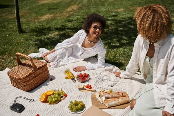 Молоді афроамериканські подружки розмовляють на ковдрі біля фруктів під час літнього пікніка — стокове фото