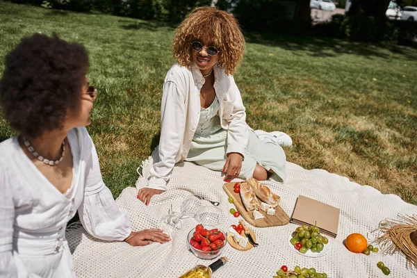Novias afroamericanas jóvenes en vestidos blancos relajándose en el picnic de verano cerca de frutas y vino - foto de stock