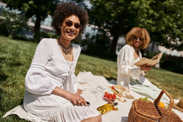 Mujer afroamericana con teléfono inteligente mirando a la cámara cerca de la novia, picnic de verano - foto de stock
