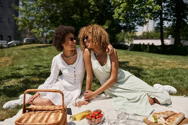 Happy african american girlfriends looking at each other, blanket, food and wine, summer picnic — Stock Photo