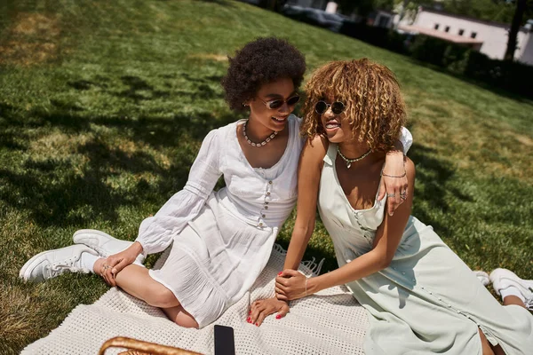Felizes namoradas africanas americanas em óculos de sol elegantes abraçando no cobertor no parque, piquenique de verão — Fotografia de Stock