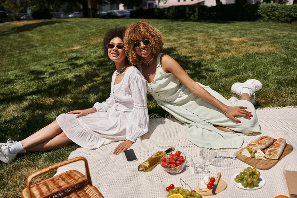 Elegantes novias afroamericanas descansando en manta cerca de la comida y el vino en el picnic de verano - foto de stock
