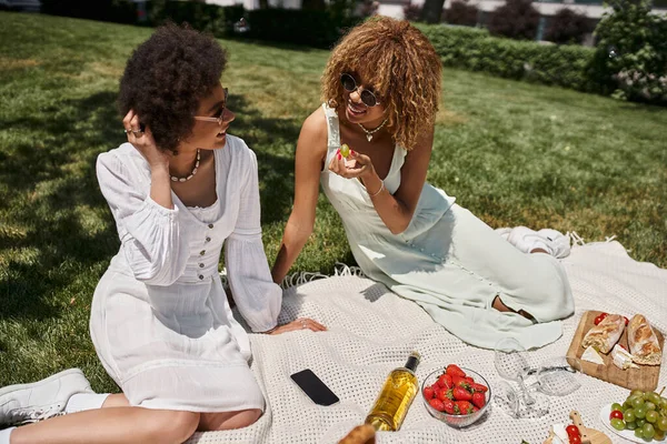 Афроамериканські подружки в сонцезахисних окулярах їдять фрукти і розмовляють під час пікніка — стокове фото